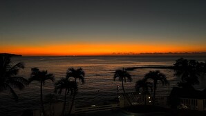 Une vue sur un coucher de soleil au-dessus d’une étendue d’eau, avec des palmiers et un bâtiment au premier plan.
