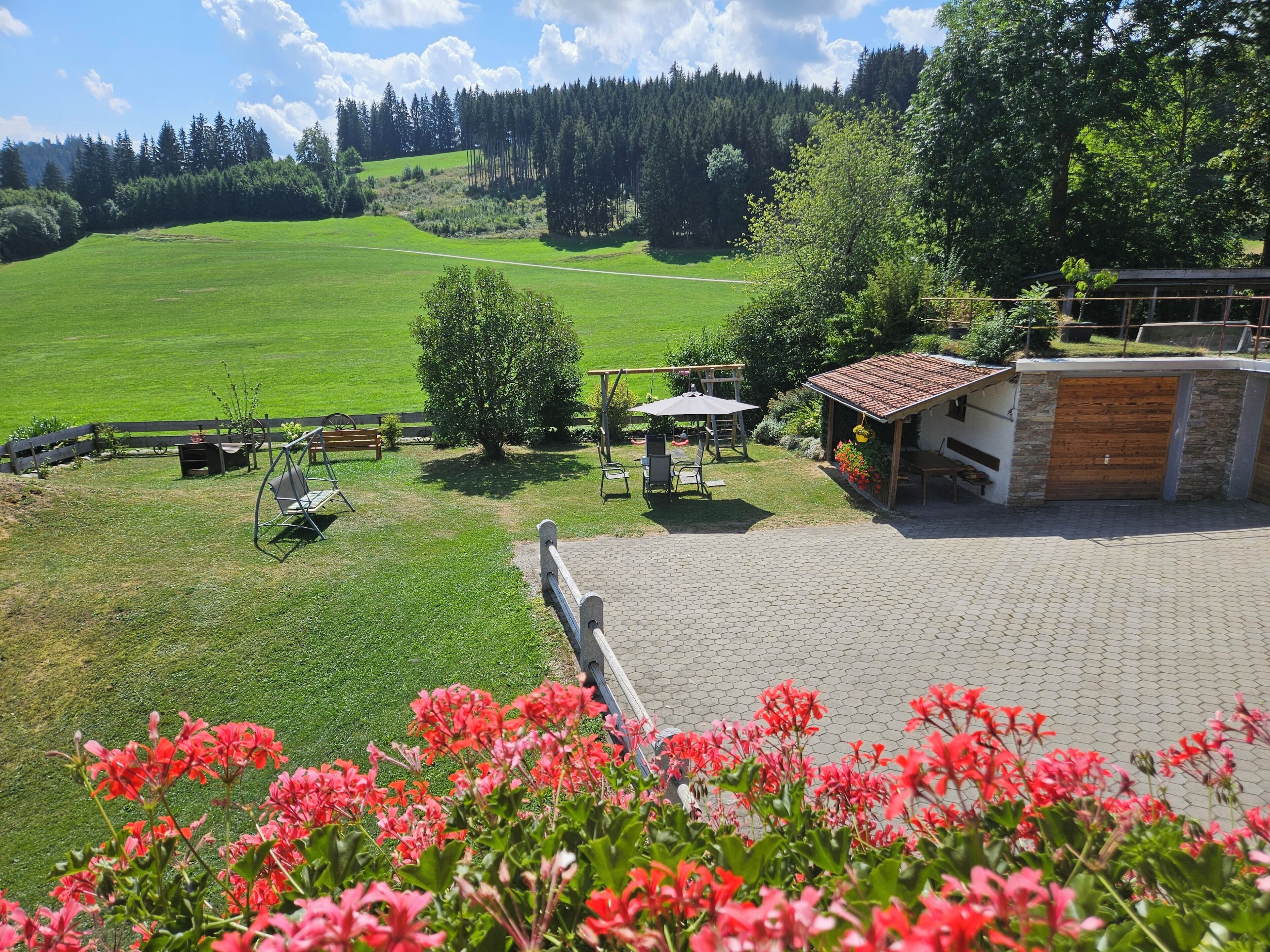 A patio area with a table and chairs, a small garden with red flowers, and a view of a green field and trees in the background.