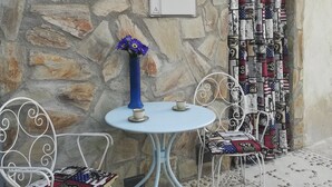 Een stenen muur met een houten bord waarop “LE CORRALE” staat. Er is een kleine tafel met twee stoelen, een vaas met bloemen en een decoratief muurpaneel met diverse objecten.