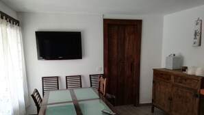 Een eethoek met een tafel met glazen blad, houten stoelen, een aan de muur bevestigde televisie en een houten kast.