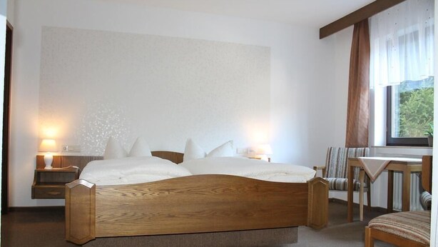 A bedroom with a wooden bed, two bedside tables with lamps, a chair, and a window with curtains. - Quite double rooms with breakfast (Hornberg)
