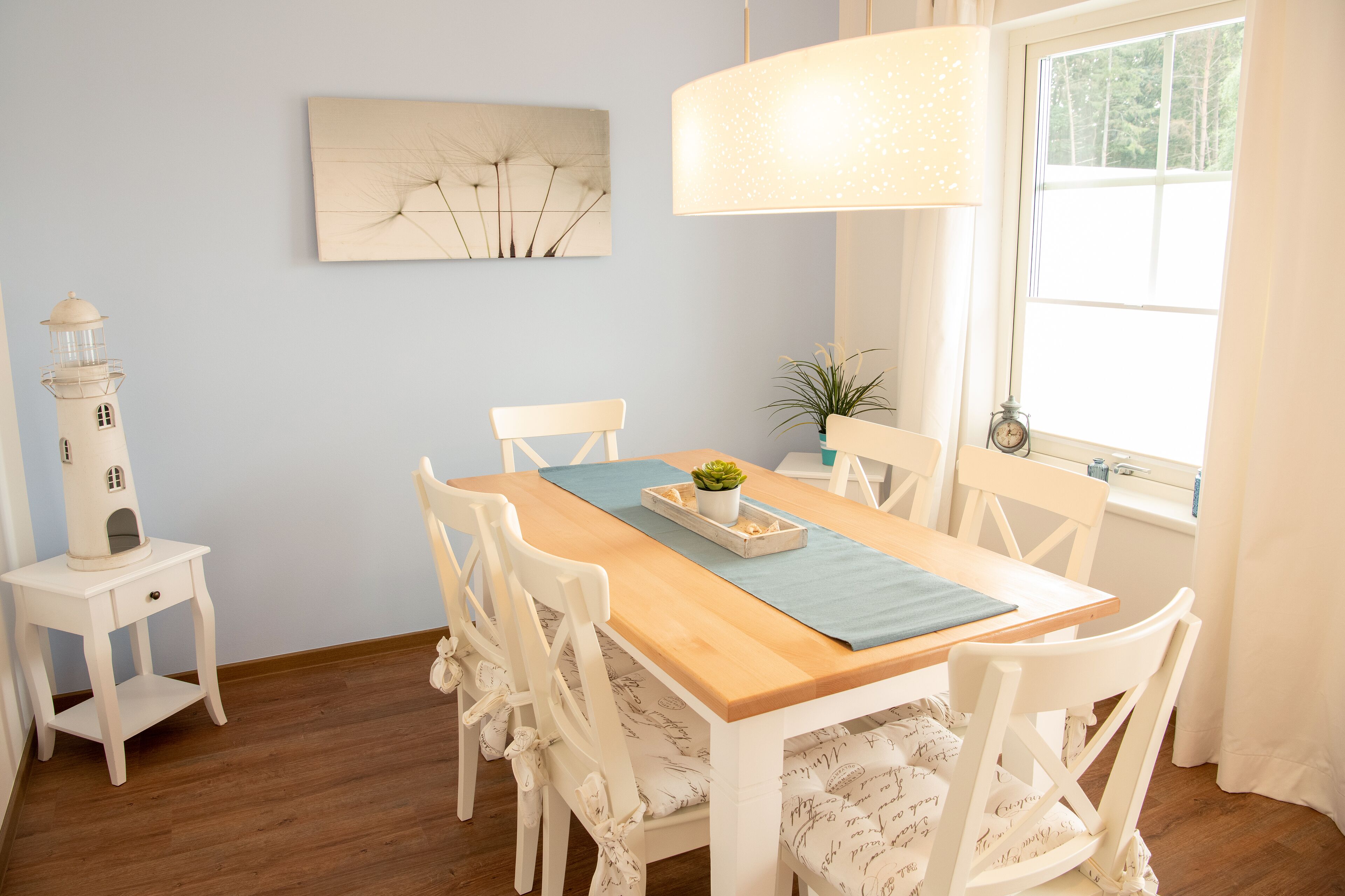 Een eetruimte met een houten tafel, witte stoelen, een klein bijzettafeltje met een model van een vuurtoren, een wanddecoratie en een raam met gordijnen.