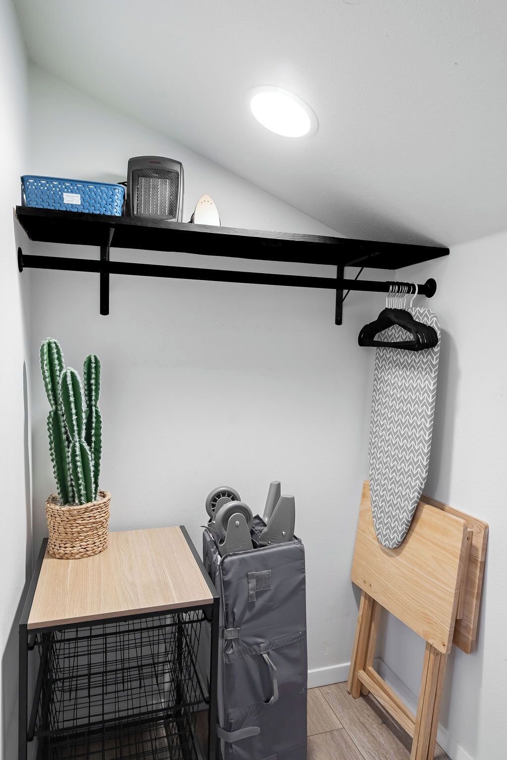 A small kitchenette with a wooden table, a hanging rack, a cactus, and a portable ironing board.