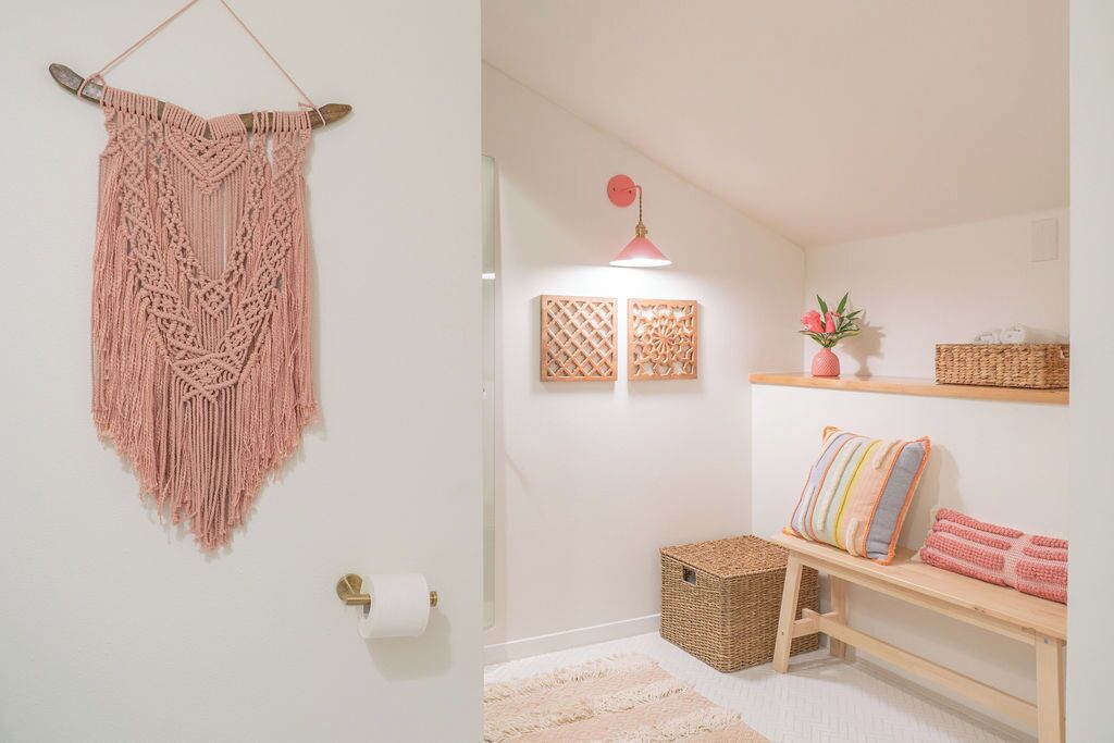 A room with a macrame wall hanging, a pink pendant light, a woven basket, a shelf with decorative pillows, and a small plant.