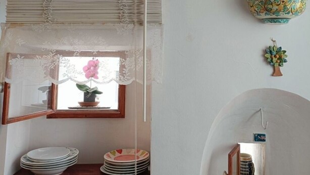 Una cocina con vigas de madera, una ventana con cortinas de encaje y una estantería con platos.