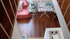 Una habitación con piso de madera, un sofá pequeño rosa, una mesa con mantel blanco y un mueble blanco con un lavamanos.