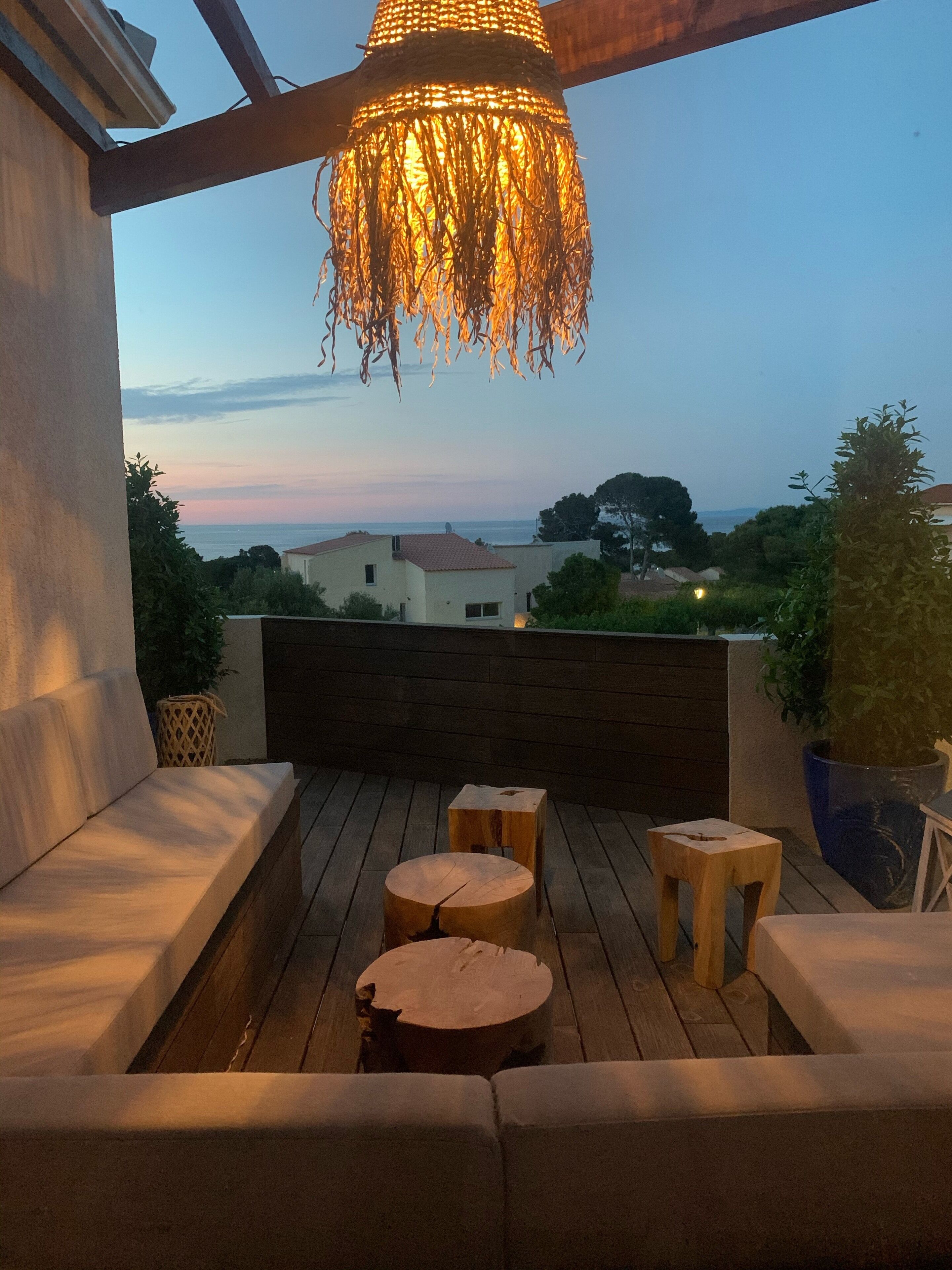 An outdoor deck with wooden furniture, a hanging light fixture, and a view of the sea and buildings in the background.