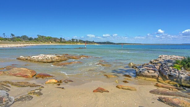 A beach with clear water, rocks, and a distant view of a lighthouse.