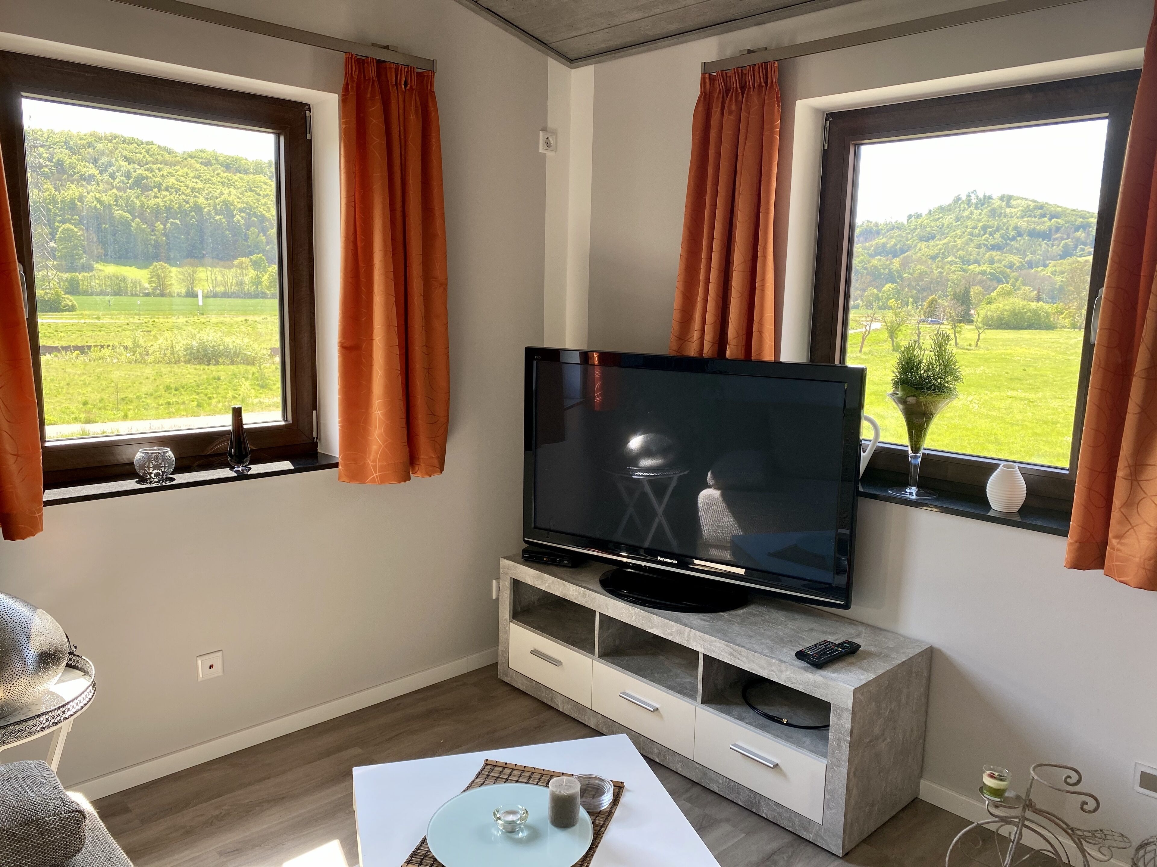 Una sala de estar moderna con un televisor de pantalla plana sobre una consola de piedra, ventanas amplias con cortinas, una mesa de comedor con un florero y vistas a un paisaje verde.