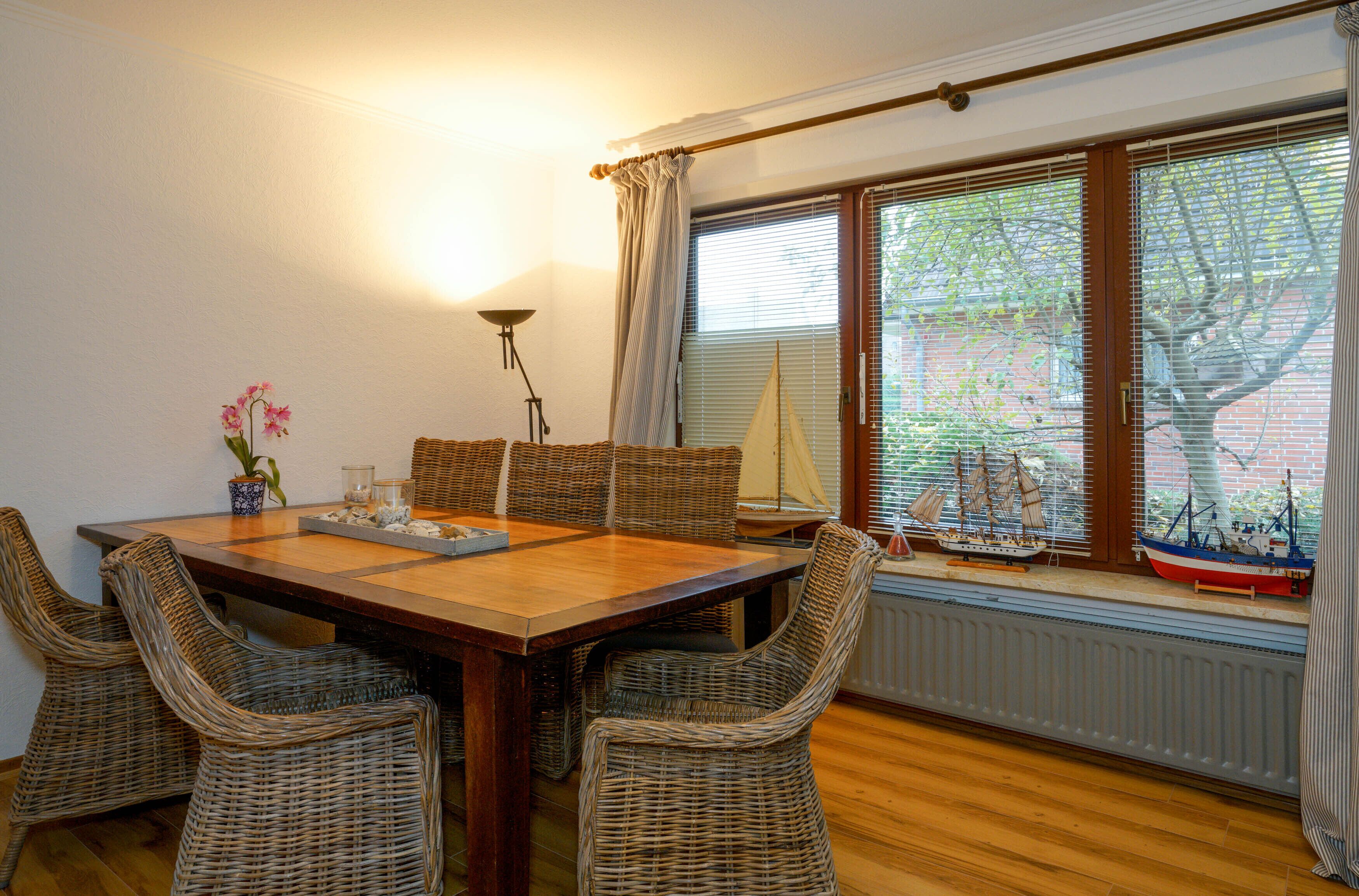 A dining area with a wooden table, wicker chairs, a vase with flowers, and a window with blinds and a sailboat model.