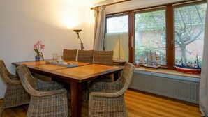 Een eethoek met een houten tafel, rieten stoelen, een vaas met bloemen, en een raam met luiken en een model zeilboot.