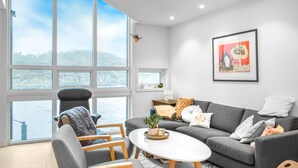 Una sala de estar moderna con un sofá gris, una silla de madera, una mesa de centro blanca y vistas a un lago a través de amplios ventanales.