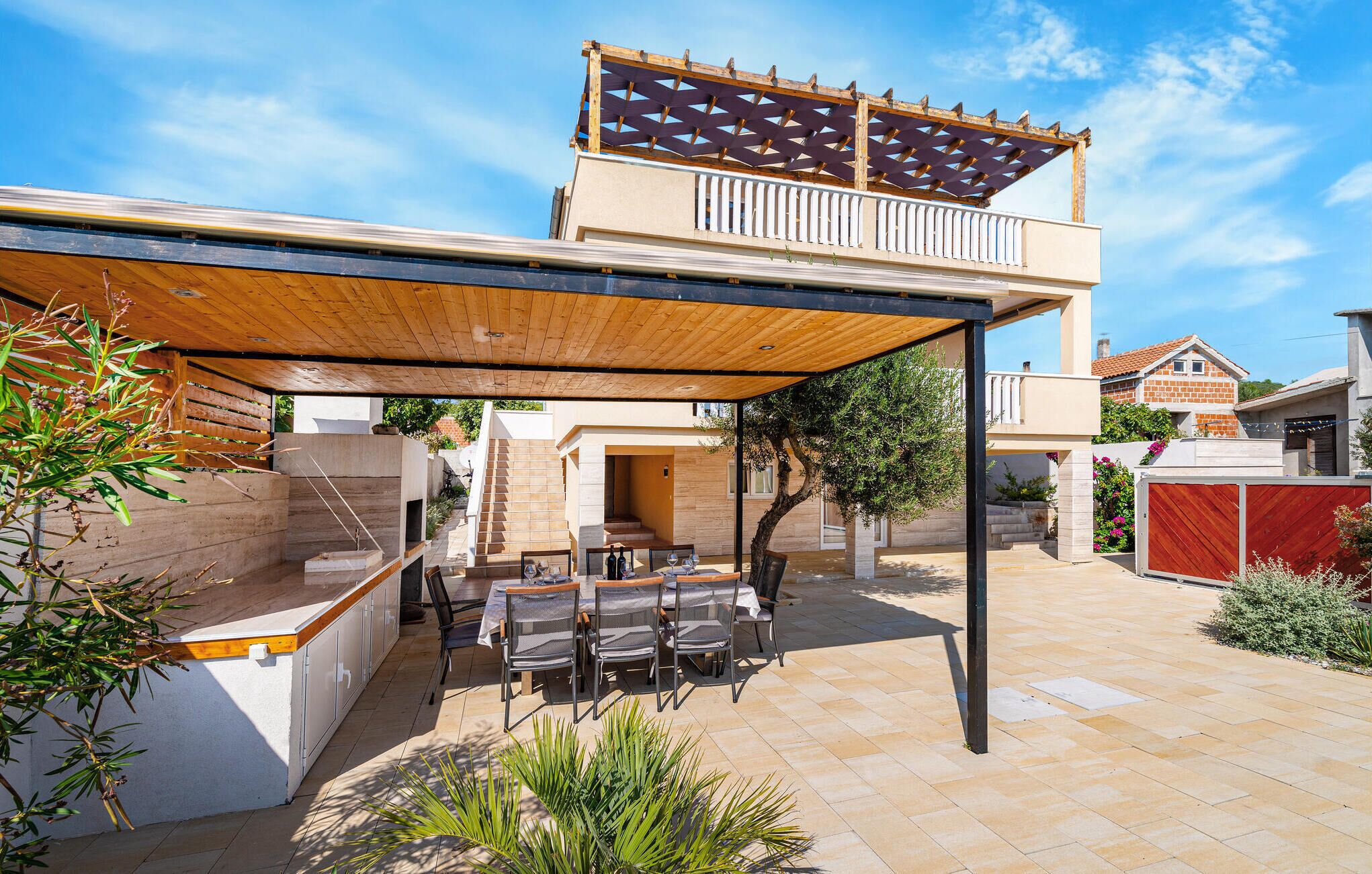 Un patio exterior moderno con una pérgola de madera, una zona de comedor con sillas y una mesa, y una escalera que lleva a un segundo piso.