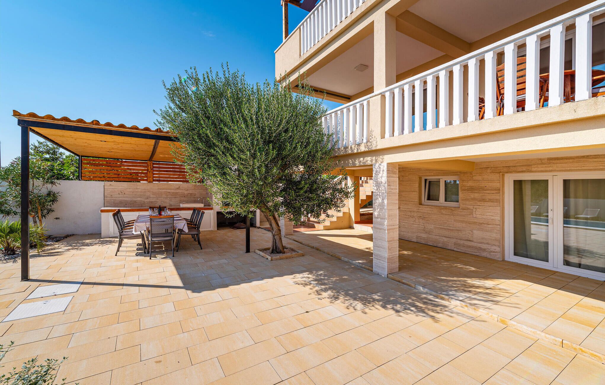A covered outdoor patio with a dining table and chairs, a small tree, and a staircase leading to an upper floor.