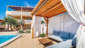 Area piscina con una pergola in legno, una piscina e un salotto con cuscini.