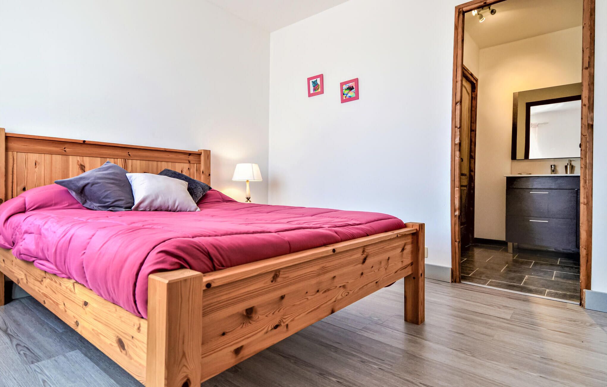 A wooden bed with a pink bedspread and pillows, a bedside lamp, and a small mirror above a dark wooden dresser.
