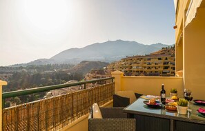 A balcony with a table set for dining, overlooking a mountainous landscape with buildings in the distance.