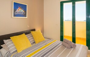 A hotel room with a bed, yellow pillows, a striped bedspread, a framed artwork on the wall, and a green door leading to a balcony.