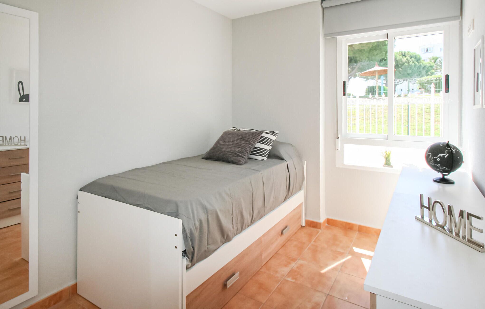 A small, neatly arranged bedroom with a single bed, a small desk with a globe, and a window with a view of a garden and buildings.