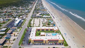 Ein Luftbild eines Küstenhotels mit langer Strandpromenade, mehreren Parkplätzen und einem zentralen Gebäude mit Schwimmbad.