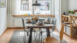 Uma área de refeições com uma mesa posta para quatro pessoas, uma janela com persianas, um radiador e uma estante com livros e uma planta.