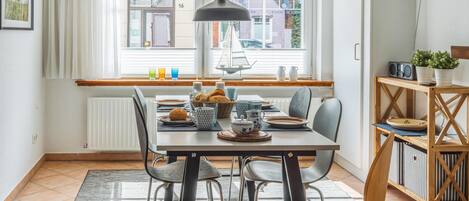 A dining area with a table set for four, a window with blinds, a radiator, and a bookshelf with books and a plant.