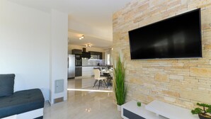 Una sala moderna con una televisión de pantalla plana montada en una pared de piedra, un sofá, un comedor con mesa y sillas, y una cocina al fondo.