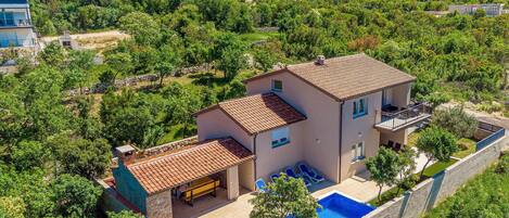 A two-story house with a swimming pool, surrounded by lush greenery and a stone wall.
