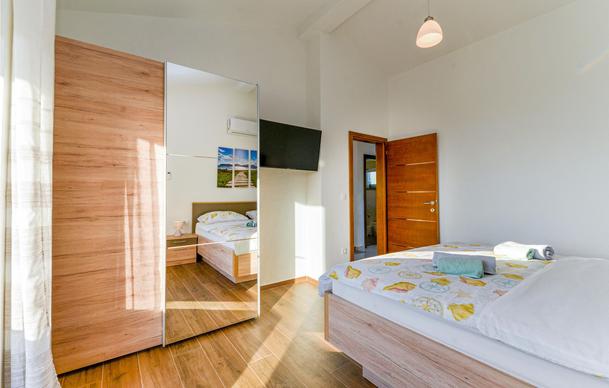 A bedroom with a wooden wardrobe, a bed with a colorful duvet, a bedside table, and a mirror.