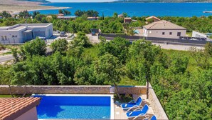 Zona de piscina con agua azul, rodeada de vegetación y con vistas a un paisaje costero con edificios y montañas.