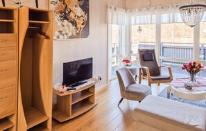 Ein modernes Wohnzimmer mit Fernseher, Holzmöbeln, einem Sessel, einem Tisch mit einer Vase und Blick auf einen Balkon.