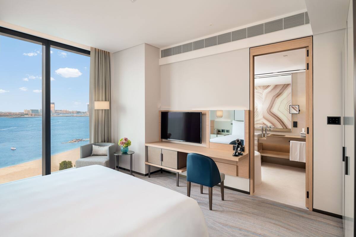 A hotel room with a large bed, a desk with a TV, a view of the sea, and a bathroom with a mirror and sink.