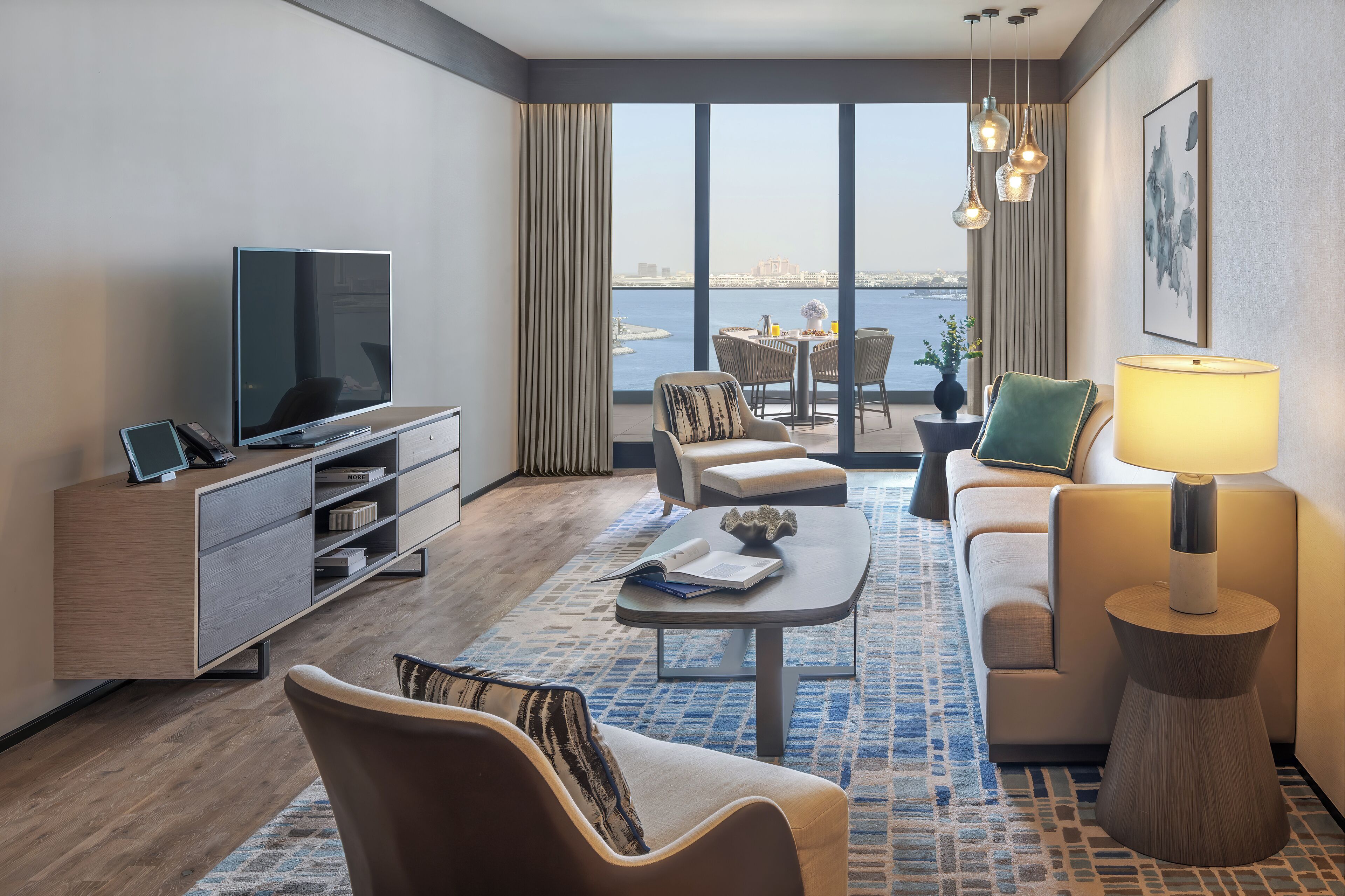 Una habitación de hotel moderna con un televisor grande, asientos cómodos, un comedor con vista al agua y una lámpara sobre una mesita.
