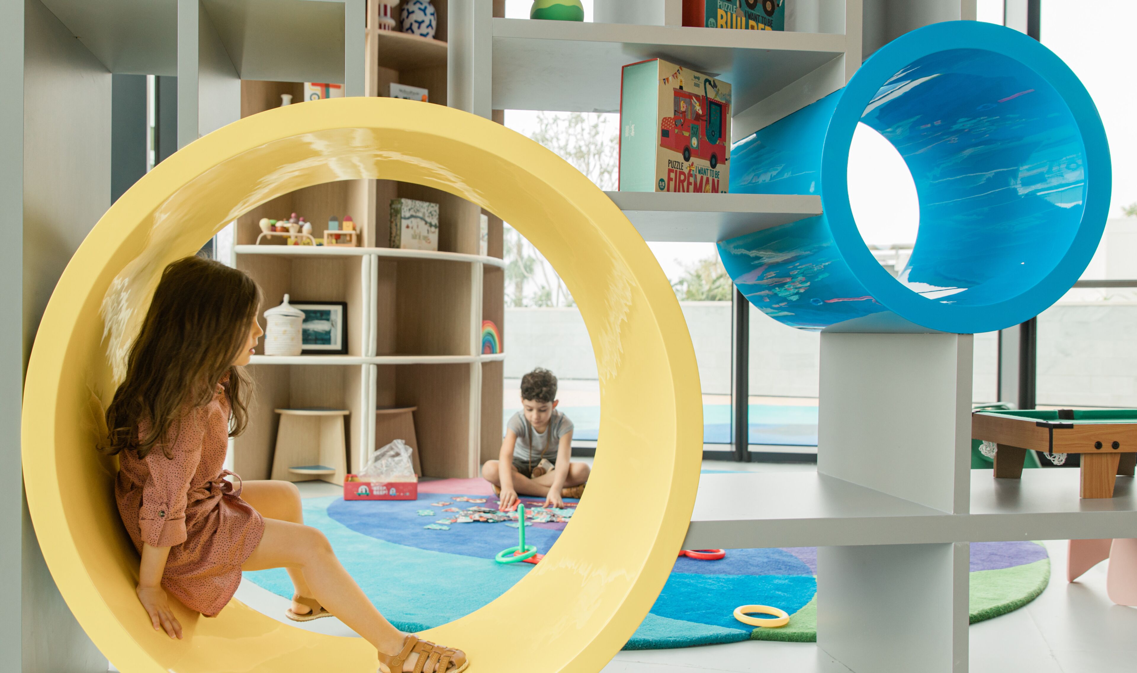 A child sitting in a yellow circular play structure with a blue slide, surrounded by shelves with toys and books.