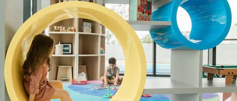 A child sitting in a yellow circular play structure with a blue slide, surrounded by shelves with toys and books.