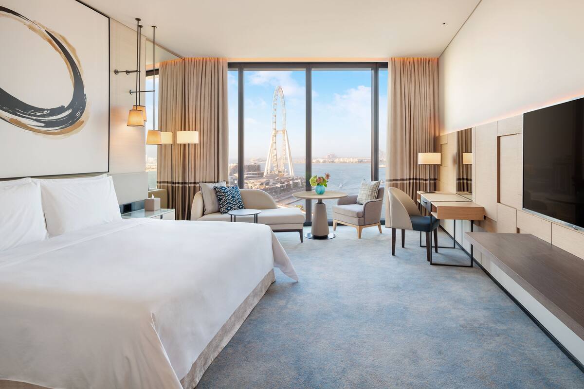 A hotel room with a large bed, a desk, two chairs, a TV, and a view of a Ferris wheel.