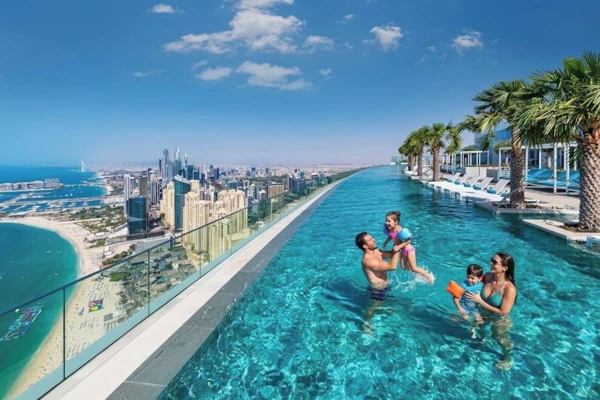 A poolside scene with a city view, palm trees, and a beach.
