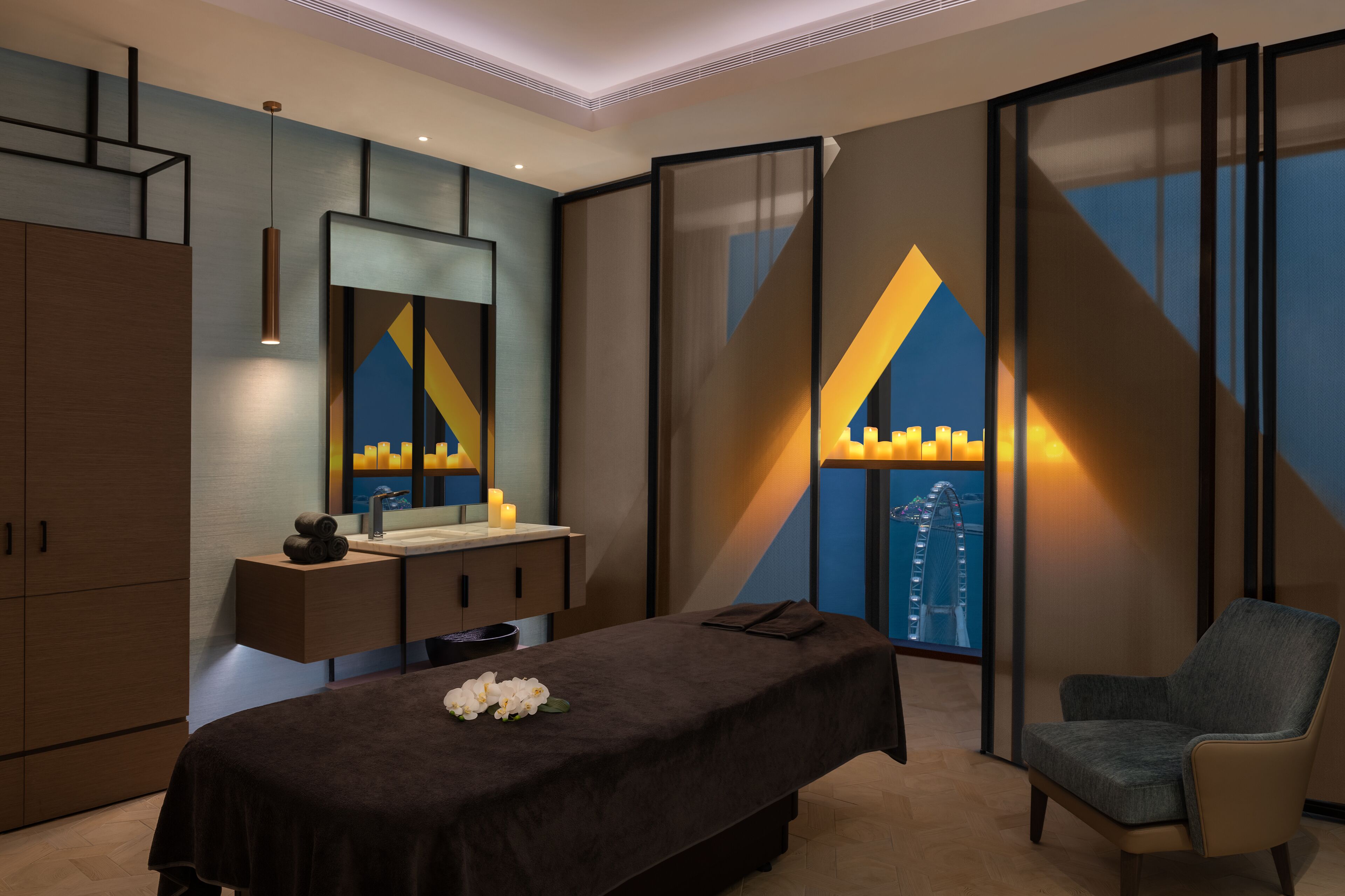 A modern hotel room with a bed, a massage table, a chair, a vanity with a mirror, and a large window with a view of a Ferris wheel.