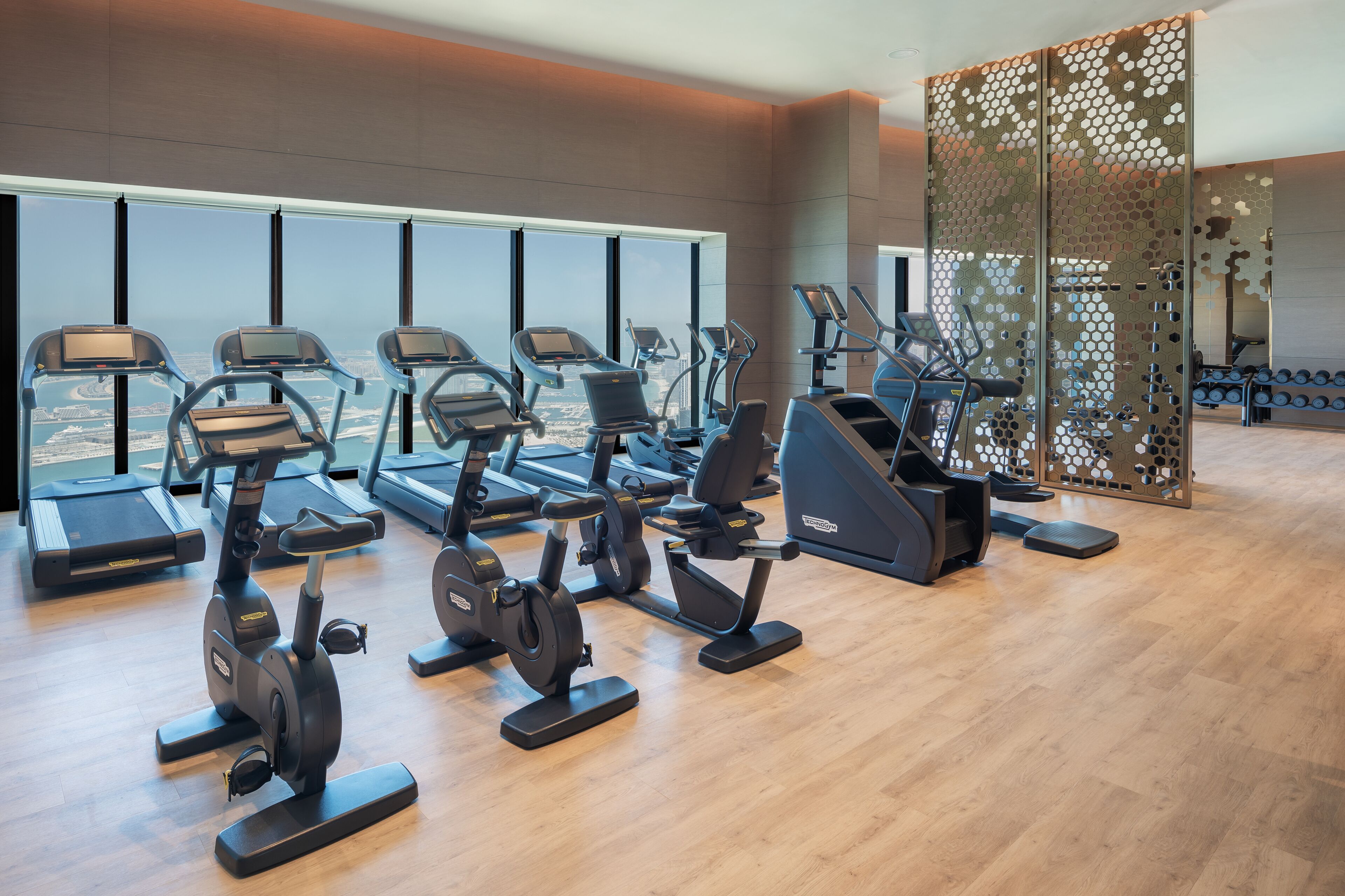 A gym with various exercise machines, large windows, and a view of a cityscape.