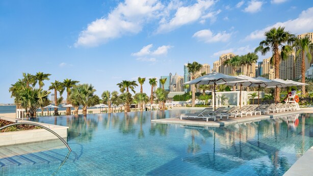 A swimming pool with lounge chairs and umbrellas, surrounded by palm trees and a city skyline in the background. - Address Beach Resort (Dubai)
