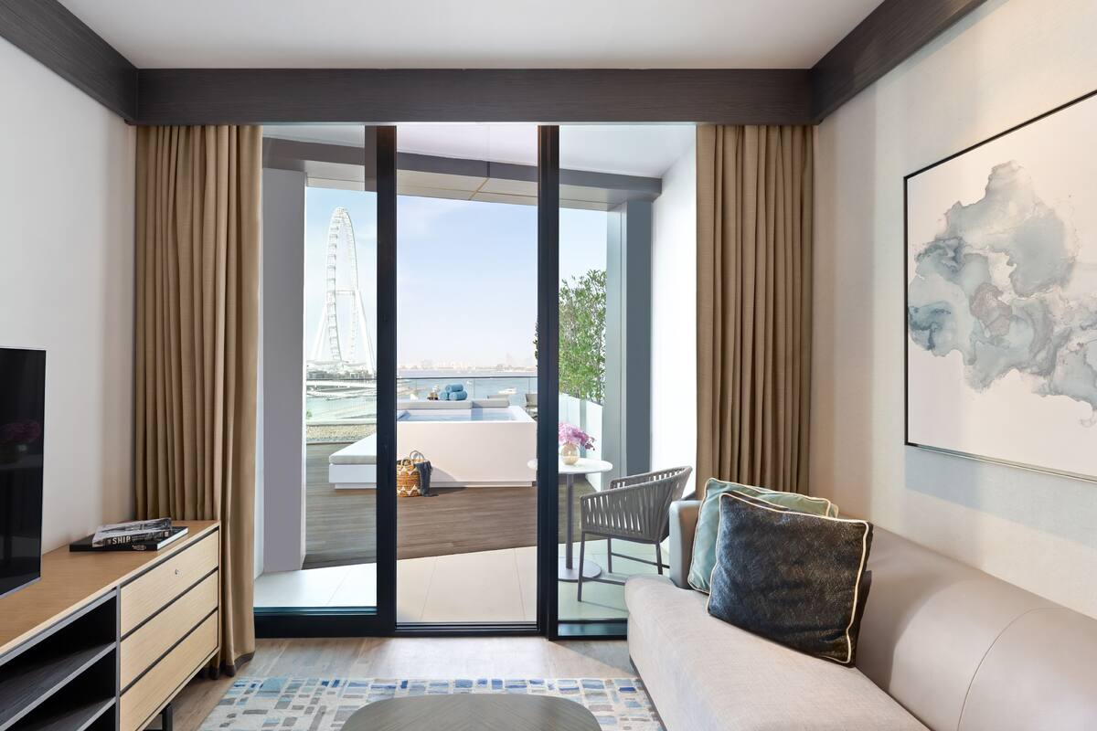 A modern hotel room with a sofa, a TV, a small table, and a view of a bridge and cityscape through the glass door.