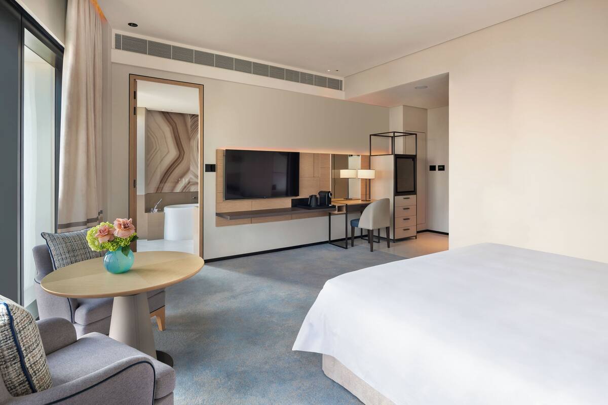 A modern hotel room with a large bed, a desk with a chair, a TV, and a small table with flowers.