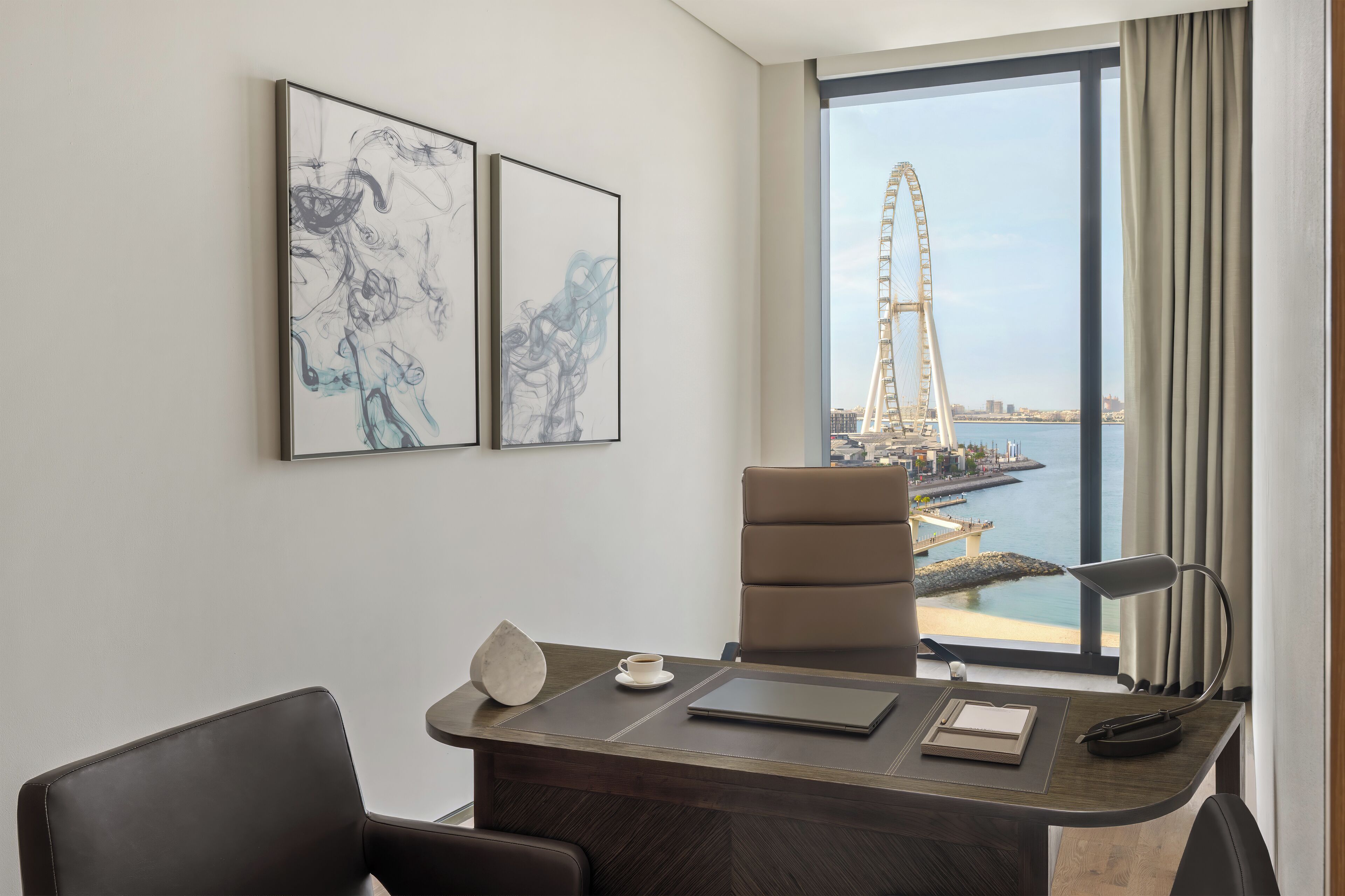 A modern hotel room with a large window offering a view of a Ferris wheel and a bridge. The room features a desk with a laptop, a chair, and a lamp.