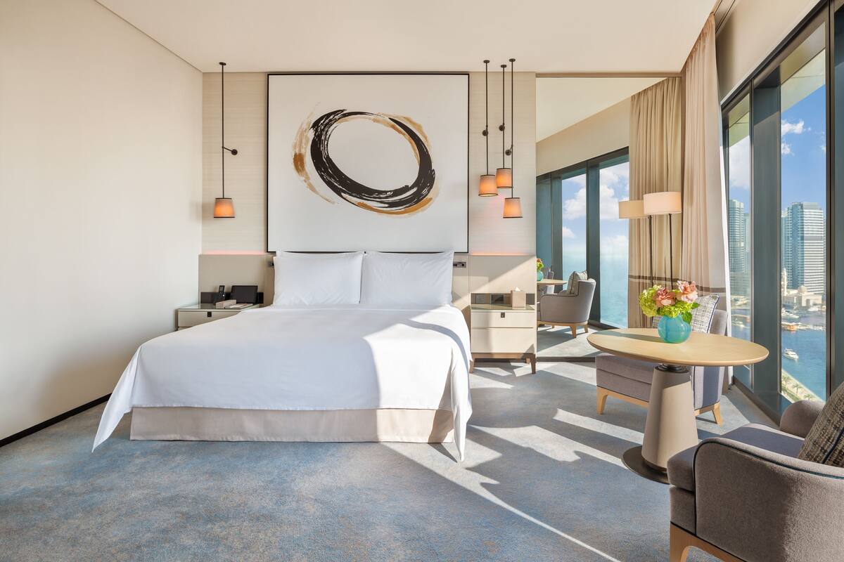 A modern hotel room with a large bed, a desk, a chair, and a view of the city.