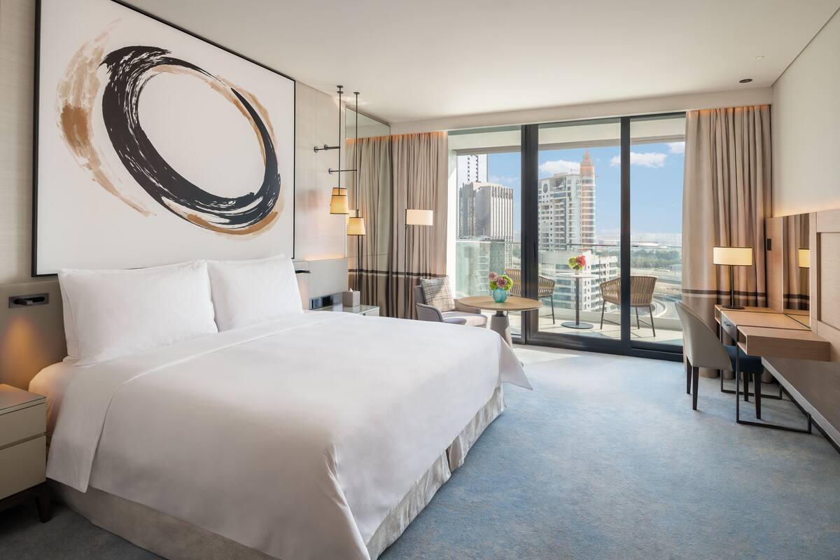 A hotel room with a large bed, a desk, and a view of the cityscape through large windows.