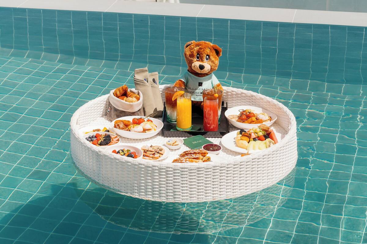 A poolside breakfast setup with a teddy bear, a white wicker table, and a view of a bridge and water.