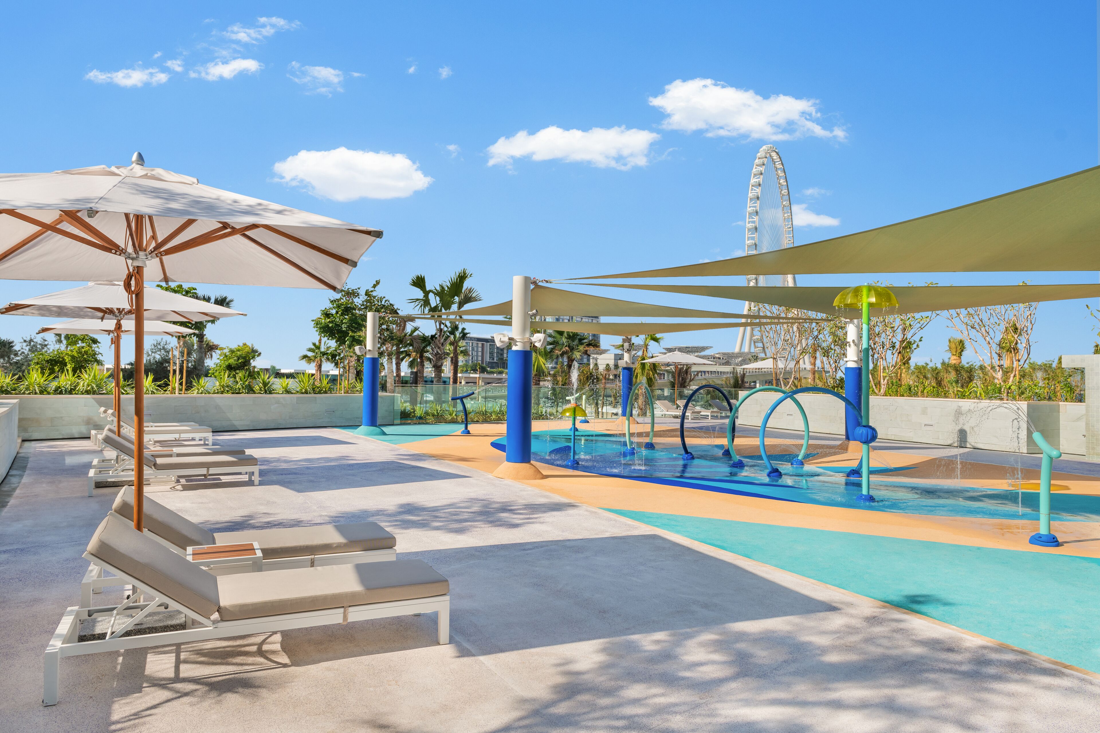A rooftop pool area with lounge chairs and umbrellas, a water feature, and a Ferris wheel in the background.
