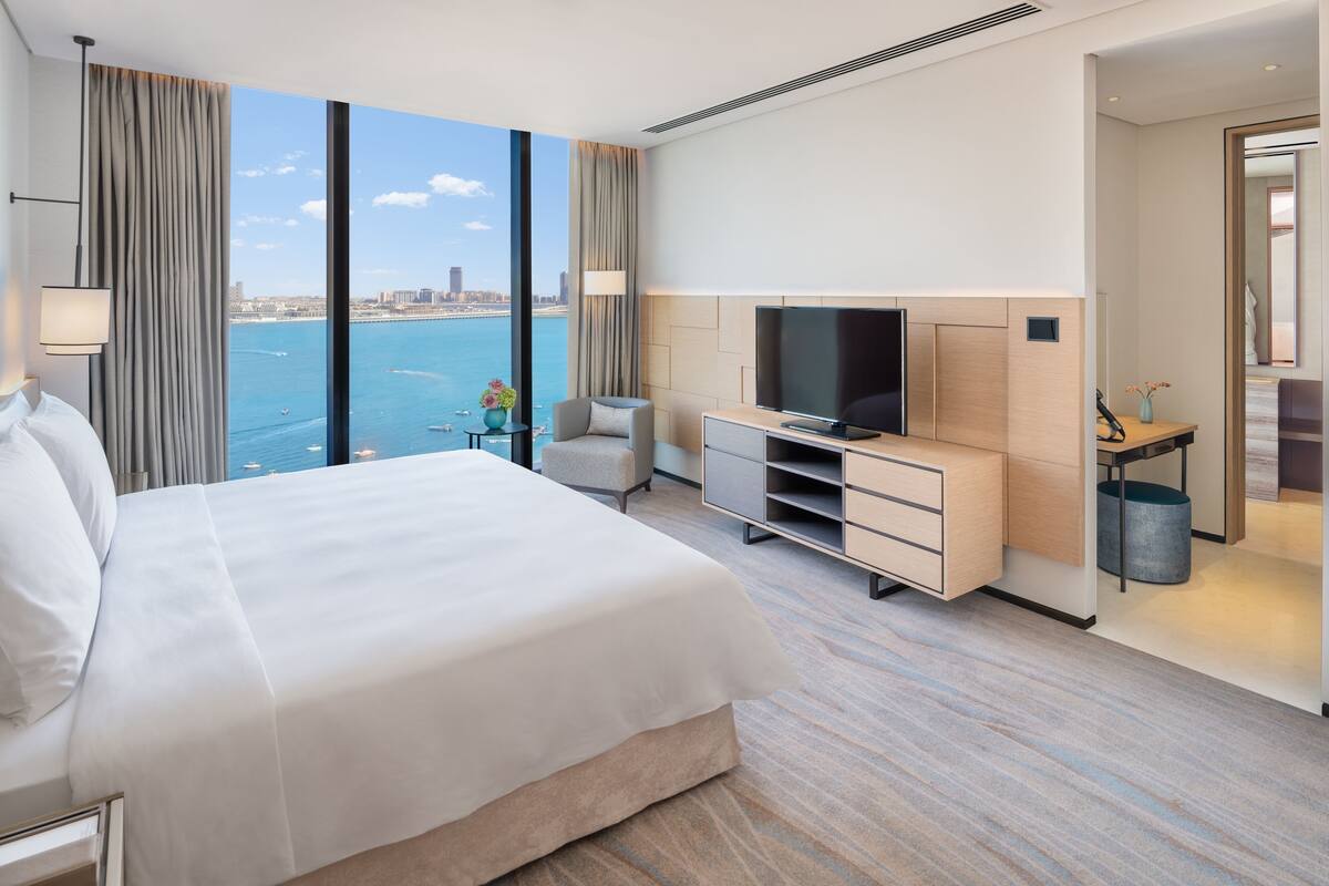 A modern hotel room with a large bed, a TV, a desk, and a view of the sea.
