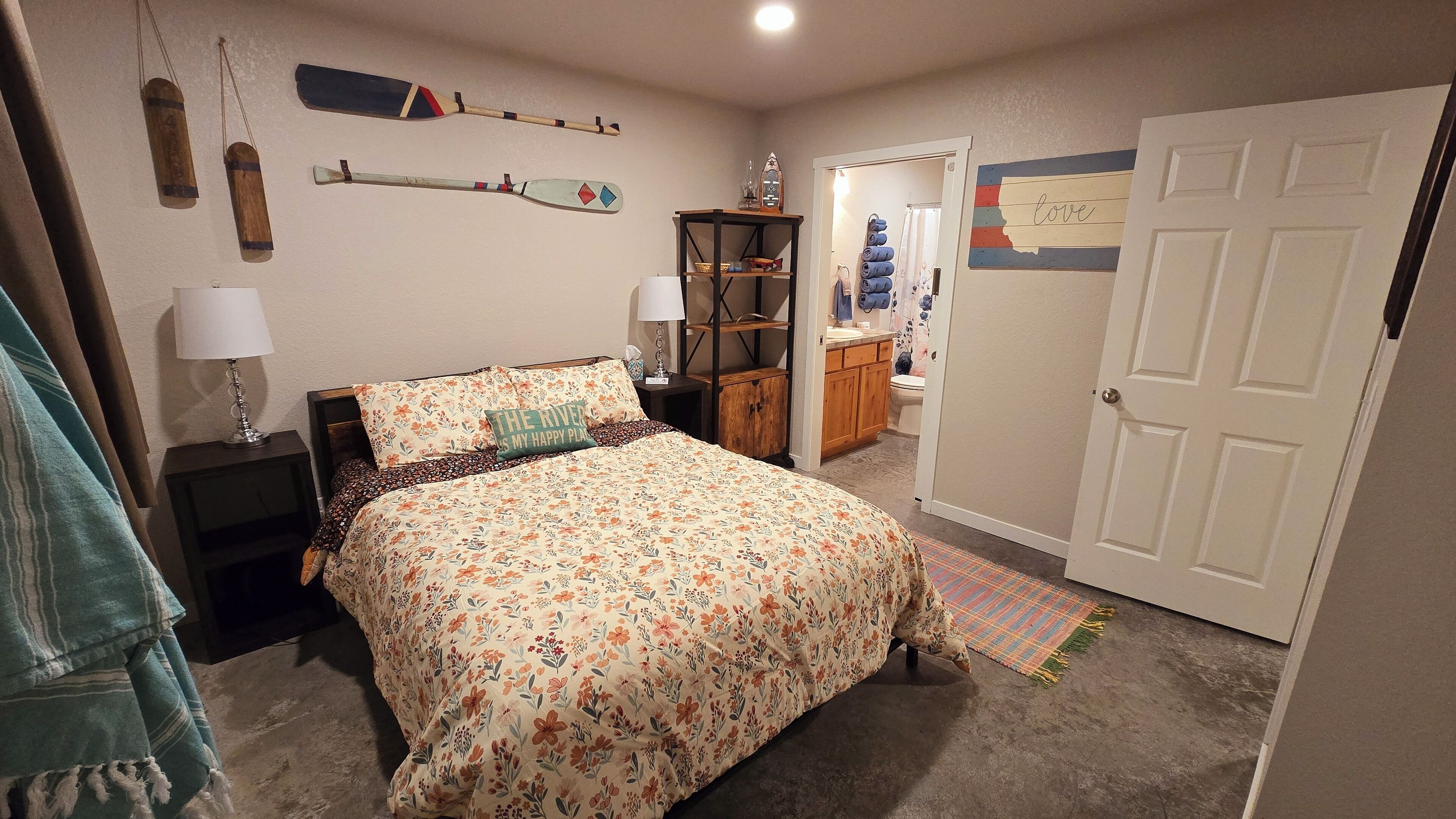 A bedroom with a floral bedspread, a bedside table with a lamp, a wooden shelf with decorative items, and a door with a 'love' sign.