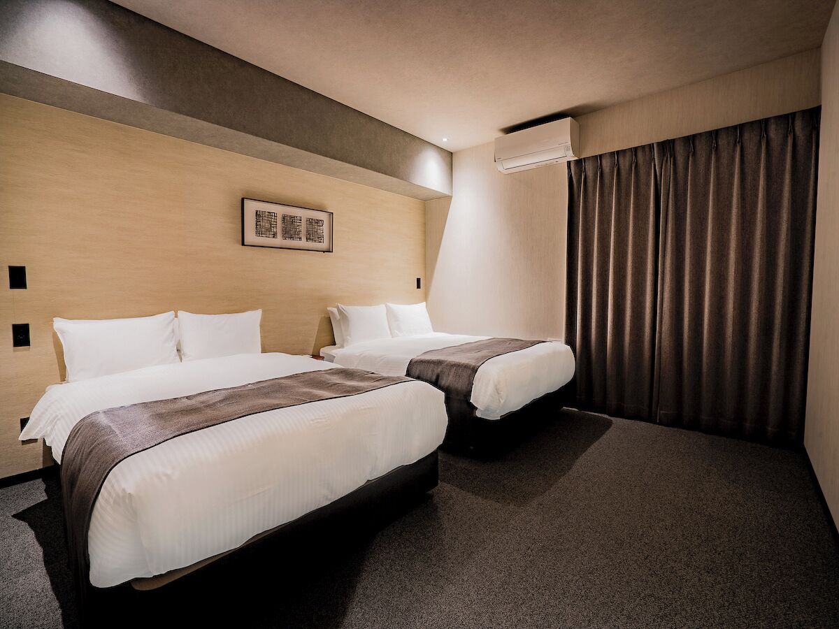 A hotel room with two beds, a large window with curtains, a wall-mounted air conditioner, and a framed picture on the wall.
